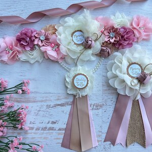 May include: A set of three sashes for a baby shower. The sashes are made of pink, beige, and brown fabric with a white flower and a gold circle with the text "Mommy To Be" or "Daddy To Be".