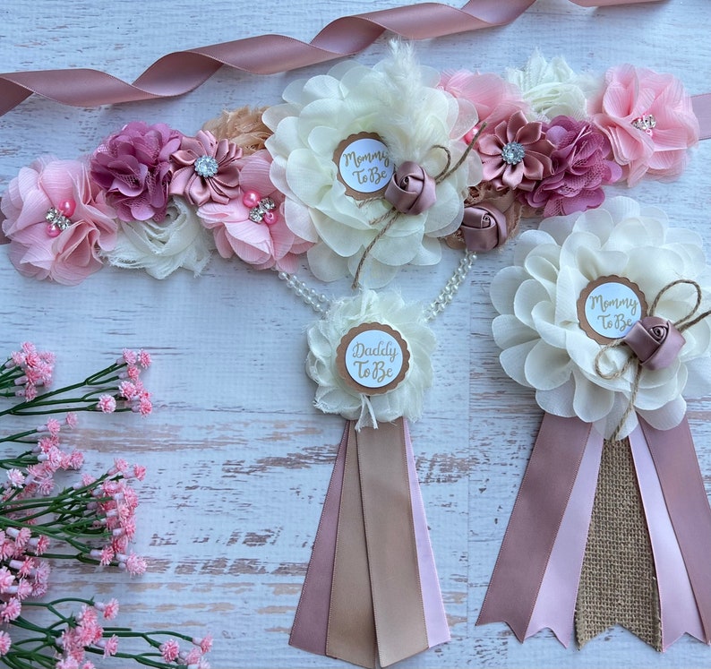 May include: A set of three sashes for a baby shower. The sashes are made of pink, beige, and brown fabric with a white flower and a gold circle with the text "Mommy To Be" or "Daddy To Be".