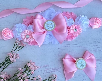 May include: Pink and white satin sash with a large pink bow and a smaller pink bow. The sash has a white lace trim and two gold glitter circles with the text "Mommy To Be" and "Daddy To Be".