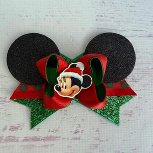 May include: A red and green Christmas bow with a Mickey Mouse character wearing a Santa hat in the centre. The bow is made of glitter fabric and has black felt Mickey Mouse ears on either side.