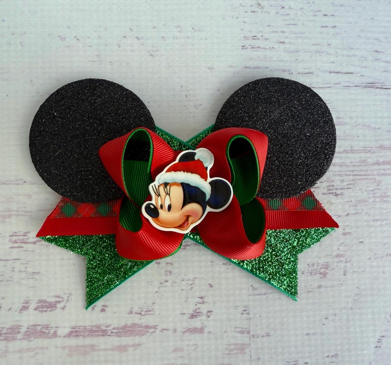 May include: A red and green Christmas bow with a Mickey Mouse character wearing a Santa hat in the centre. The bow is made of glitter fabric and has black felt Mickey Mouse ears on either side.