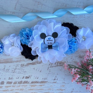 May include: A blue and white satin sash with a white flower and a black and white Mickey Mouse design. The sash has a blue bow and a blue and white flower. The sash has a tag that says "Mommy To Be".