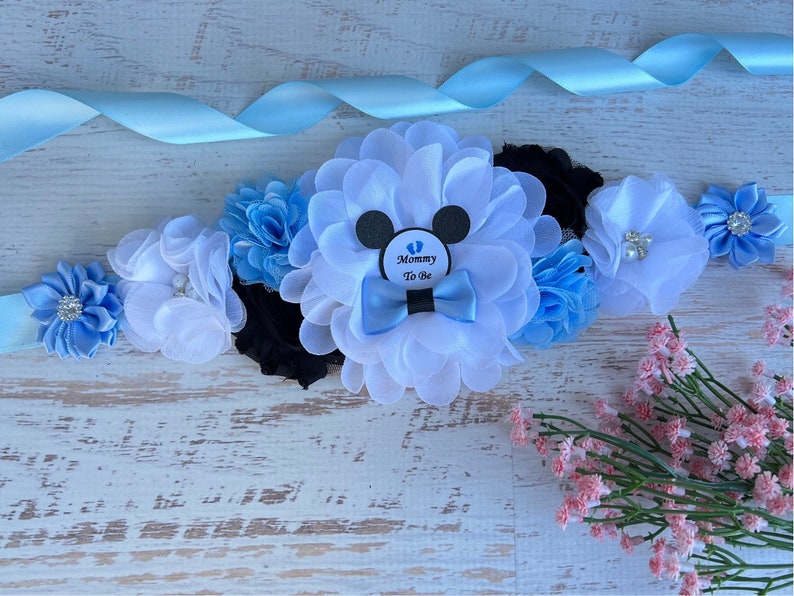 May include: A blue and white satin sash with a white flower and a black and white Mickey Mouse design. The sash has a blue bow and a blue and white flower. The sash has a tag that says "Mommy To Be".