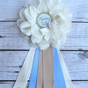 K&ouml;nnte beinhalten: Wei&szlig;e Blume mit einem goldenen Glitzerkreis, auf dem "Mommy To Be" steht, und einem wei&szlig;en Satinband. Die Blume ist an einem wei&szlig;en, blauen und braunen Band befestigt.