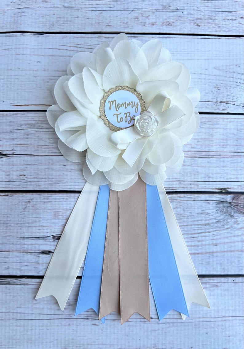K&ouml;nnte beinhalten: Wei&szlig;e Blume mit einem goldenen Glitzerkreis, auf dem "Mommy To Be" steht, und einem wei&szlig;en Satinband. Die Blume ist an einem wei&szlig;en, blauen und braunen Band befestigt.
