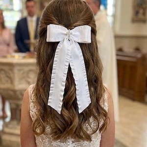 May include: A white satin hair bow with a rhinestone cross embellishment, worn in the hair. The bow is paired with a white lace dress. The person's hair is styled in loose waves.