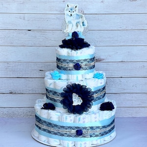 May include: A three-tiered diaper cake decorated with blue and white patterned diapers, black and silver ribbon, and blue and black flowers. A white wolf figurine sits on top of the cake.