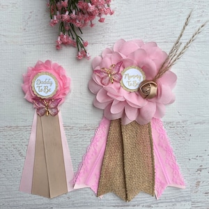May include: Two pink and gold baby shower sashes with the text "Mommy to Be" and "Daddy to Be". Each sash features a pink flower, a gold butterfly, and a pink and brown ribbon.