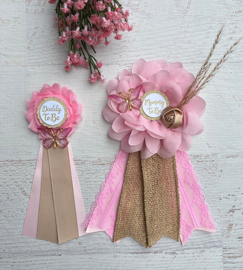May include: Two pink and gold baby shower sashes with the text "Mommy to Be" and "Daddy to Be". Each sash features a pink flower, a gold butterfly, and a pink and brown ribbon.