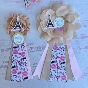 Puede incluir: Dos boutonnières de tela floral beige y rosa con una cinta blanca impresa con torres Eiffel, globos aerostáticos y bicicletas. La parte superior de cada boutonnière tiene un círculo blanco con un contorno de brillo dorado y un texto que dice "Mommy To Be" y "Daddy To Be".