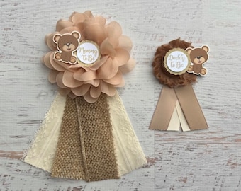 Könnte beinhalten: Zwei braune und beige Stoffschleifen mit einem braunen Teddybär und dem Text "Mommy to Be" und "Daddy to Be" auf jeder. Die Schleifen sind mit Spitze und Sackleinen verziert.