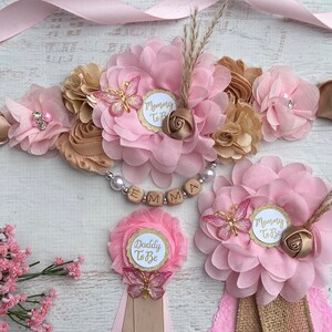 May include: Pink and gold floral sash with a wooden name tag that says "EMMA".  The sash also has a small butterfly charm and a "Mommy To Be" tag.  There are two matching pink and gold floral buttonholes with butterflies and "Mommy To Be" and "Daddy To Be" tags.