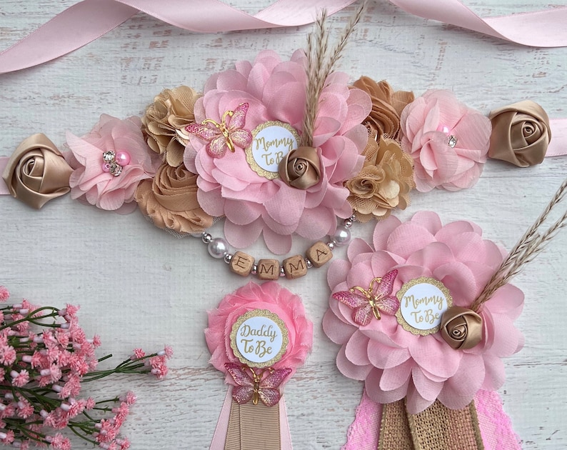 May include: Pink and gold floral sash with a wooden name tag that says "EMMA".  The sash also has a small butterfly charm and a "Mommy To Be" tag.  There are two matching pink and gold floral buttonholes with butterflies and "Mommy To Be" and "Daddy To Be" tags.