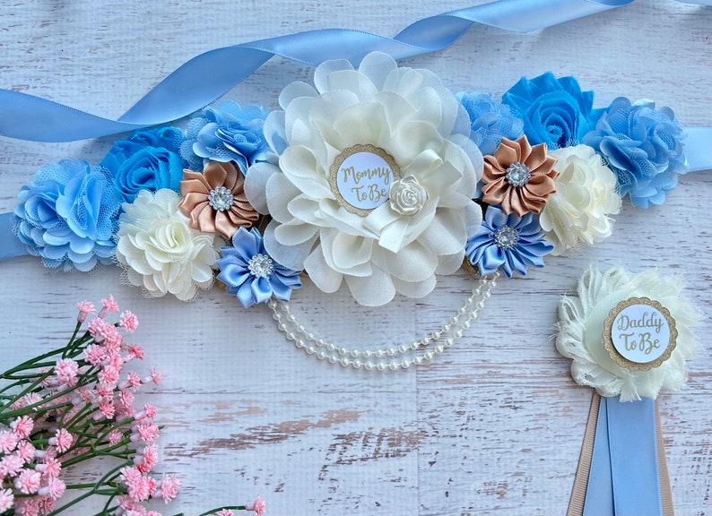 K&ouml;nnte beinhalten: Ein blaues und wei&szlig;es Schwangerschaftsband mit einer gro&szlig;en wei&szlig;en Blume und einem goldenen Glitzerkreis "Mommy to Be". Eine kleinere wei&szlig;e Blume mit einem goldenen Glitzerkreis "Daddy to Be" befindet sich auf einem blauen und braunen Band.
