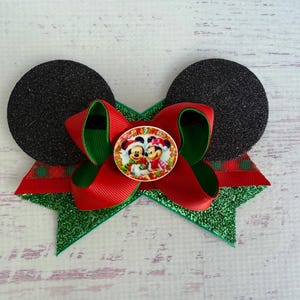 May include: Red and green Christmas bow with black glitter mouse ears and a round button featuring Mickey and Minnie Mouse wearing Santa hats.
