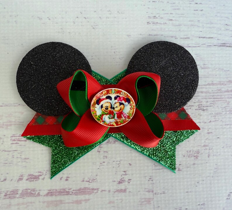 May include: Red and green Christmas bow with black glitter mouse ears and a round button featuring Mickey and Minnie Mouse wearing Santa hats.