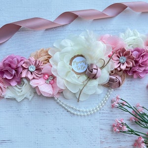May include: A blush pink, ivory, and beige floral maternity sash with a pearl strand and a "Mommy to Be" tag.