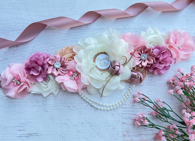 May include: A blush pink, ivory, and beige floral maternity sash with a pearl strand and a "Mommy to Be" tag.