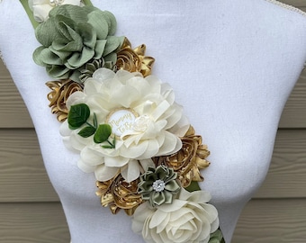 May include: A green sash with a large white and gold flower arrangement. The flowers are made of fabric and have a satin-like finish. The sash has a small white circle with the text "Mommy To Be" in gold lettering.