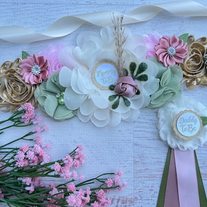 A ivory sash with a floral arrangement in shades of green, pink, and gold. The sash has a white circle with gold glitter and the text 'Mommy To Be'. There is a separate white flower with a white circle with gold glitter and the text 'Daddy To Be'.