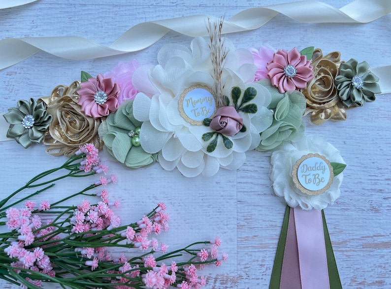 A ivory sash with a floral arrangement in shades of green, pink, and gold. The sash has a white circle with gold glitter and the text 'Mommy To Be'. There is a separate white flower with a white circle with gold glitter and the text 'Daddy To Be'.