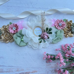 May include: A white satin sash with a floral arrangement in shades of green, pink, and gold. The flowers are made of fabric and have rhinestones in the center. The sash has a white circle with the text "Mommy To Be" in gold lettering.