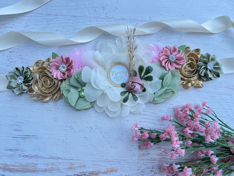 May include: A white satin sash with a floral arrangement in shades of green, pink, and gold. The flowers are made of fabric and have rhinestones in the center. The sash has a white circle with the text "Mommy To Be" in gold lettering.