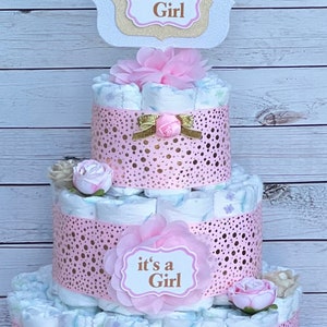 Puede incluir: Una tarta de pa&ntilde;ales de tres niveles decorada con detalles rosas y dorados. La tarta presenta una cinta rosa decorativa con lunares dorados y el texto "it's a Girl". Lazos y flores rosas y doradas adornan la tarta.