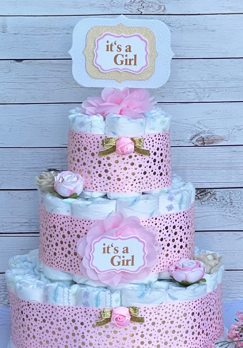 Puede incluir: Una tarta de pa&ntilde;ales de tres niveles decorada con detalles rosas y dorados. La tarta presenta una cinta rosa decorativa con lunares dorados y el texto "it's a Girl". Lazos y flores rosas y doradas adornan la tarta.
