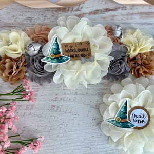 May include: A white fabric flower with a green witch hat and a brown tag that says "A Magical Bundle is on the Way" and a white fabric flower with a green witch hat and a brown tag that says "Daddy to Be".