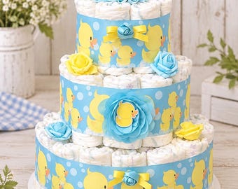 May include: A three-tiered diaper cake constructed from rolled white diapers, wrapped in light blue ribbon featuring a yellow duck pattern. The cake is decorated with yellow and blue fabric roses and topped with a yellow rubber duck.