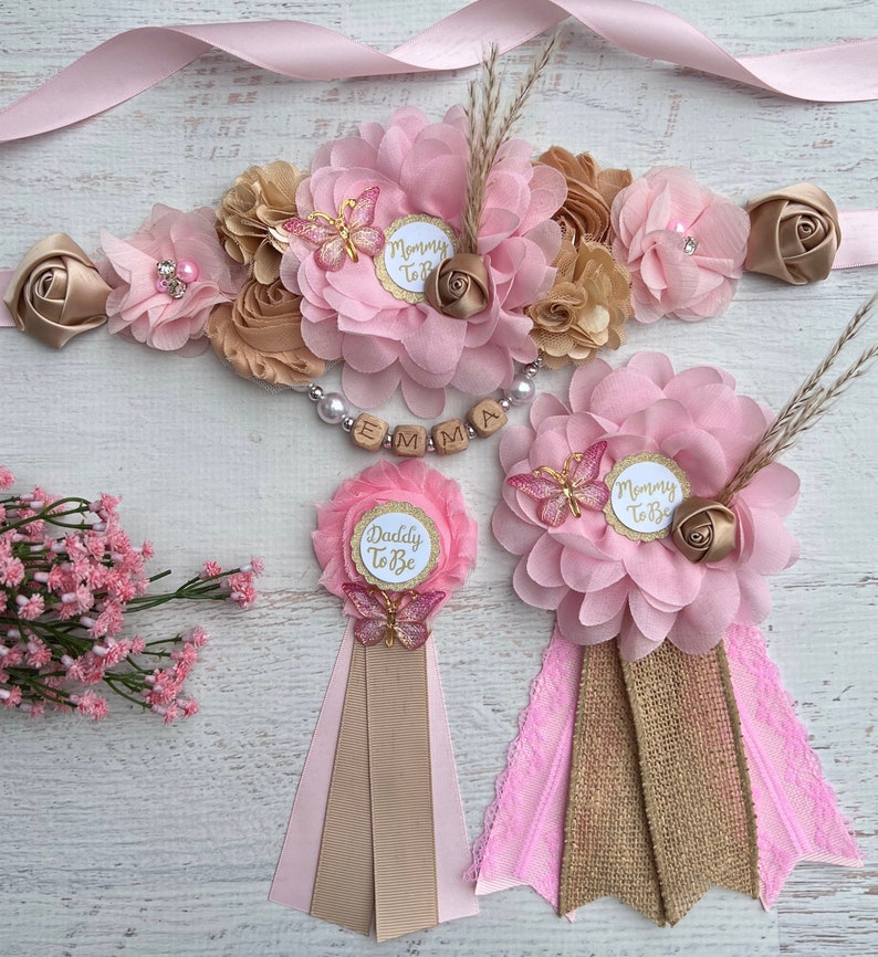 May include: Pink and gold floral sashes with the text "Mommy to Be" and "Daddy to Be" and a matching flower crown with the name "Emma" spelled out in wooden beads.