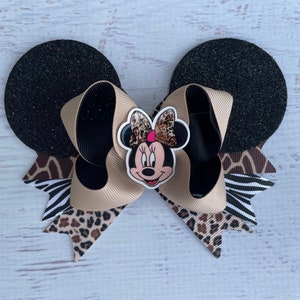 May include: A black and brown bow with a Minnie Mouse character in the centre. The bow is made of a variety of fabrics, including leopard print, zebra print, and black glitter fabric. The bow is tied with a black ribbon.