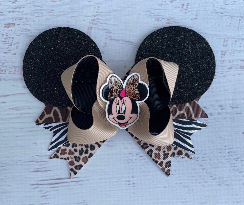 May include: A black and brown bow with a Minnie Mouse character in the centre. The bow is made of a variety of fabrics, including leopard print, zebra print, and black glitter fabric. The bow is tied with a black ribbon.