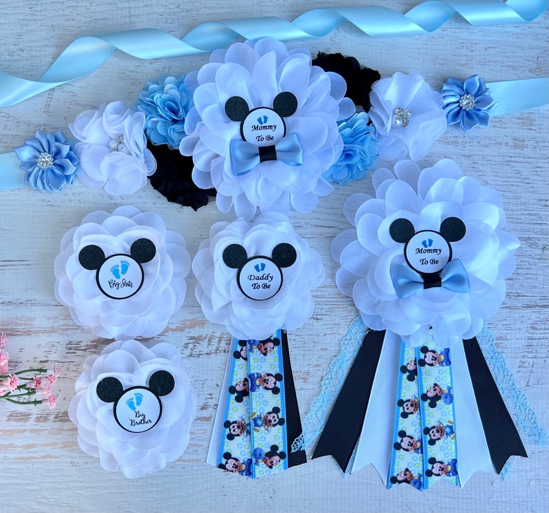 May include: A set of five blue and white Mickey Mouse themed baby shower sashes. The sashes feature a black Mickey Mouse head with a blue bow tie and the text "Mommy To Be", "Daddy To Be", and "Big Brother".