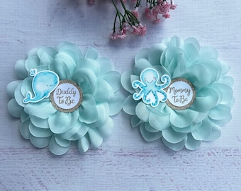 May include: Two light blue fabric flowers with a whale and an octopus on them. The whale flower has the text "Daddy To Be" and the octopus flower has the text "Mommy To Be".