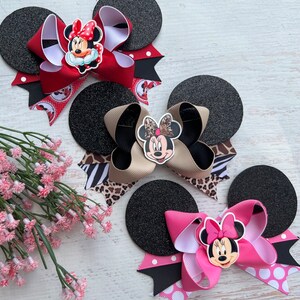 May include: Three black felt mouse ears with different coloured bows and Minnie Mouse character embellishments. The bows are red with white polka dots, tan with black and white zebra print, and pink with white polka dots.
