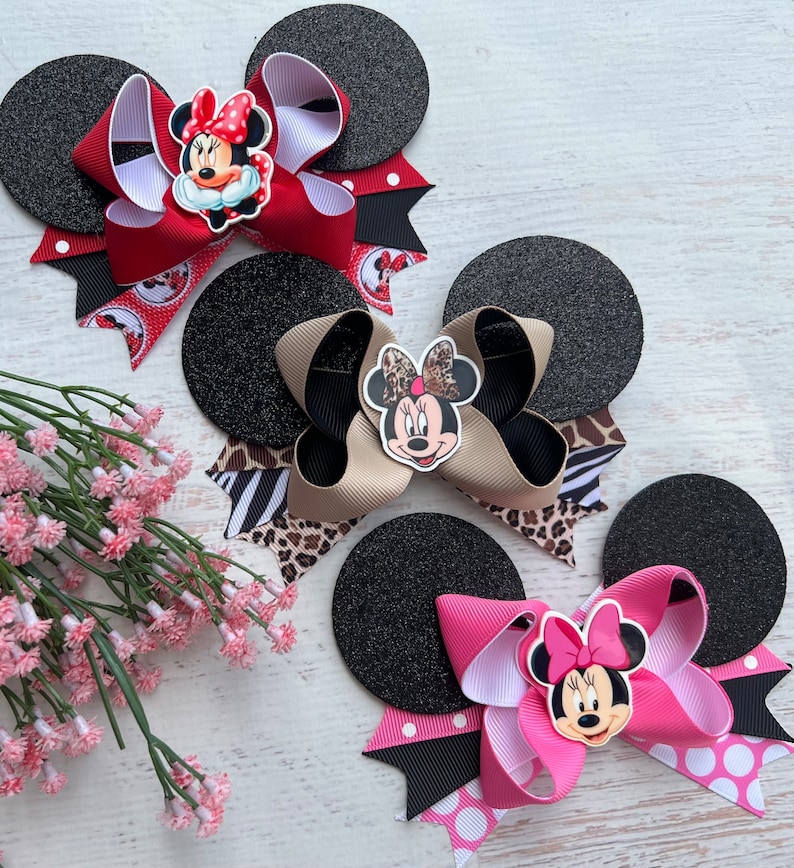 May include: Three black felt mouse ears with different coloured bows and Minnie Mouse character embellishments. The bows are red with white polka dots, tan with black and white zebra print, and pink with white polka dots.