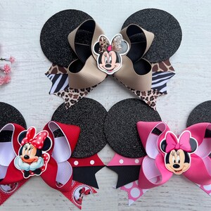 May include: Three Minnie Mouse hair bows with black glitter mouse ears. The bows are made with different coloured ribbons and feature a Minnie Mouse face in the centre. The first bow is tan, black, and white zebra print. The second bow is red, white, and black polka dot. The third bow is pink, white, and black polka dot.