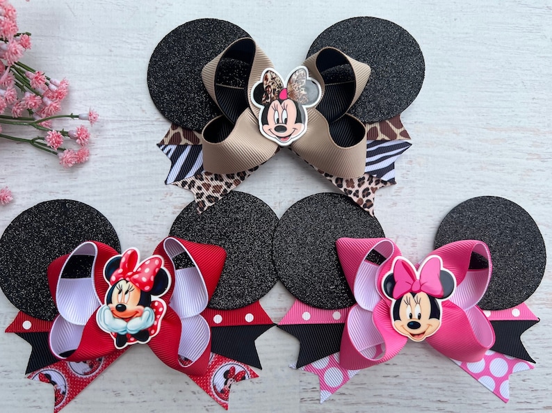 May include: Three Minnie Mouse hair bows with black glitter mouse ears. The bows are made with different coloured ribbons and feature a Minnie Mouse face in the centre. The first bow is tan, black, and white zebra print. The second bow is red, white, and black polka dot. The third bow is pink, white, and black polka dot.