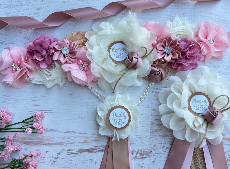 May include: A set of three floral sashes with a pink, white, and beige color scheme. The sashes feature a large flower with a circle in the center that says "Mommy to Be" or "Daddy to Be".