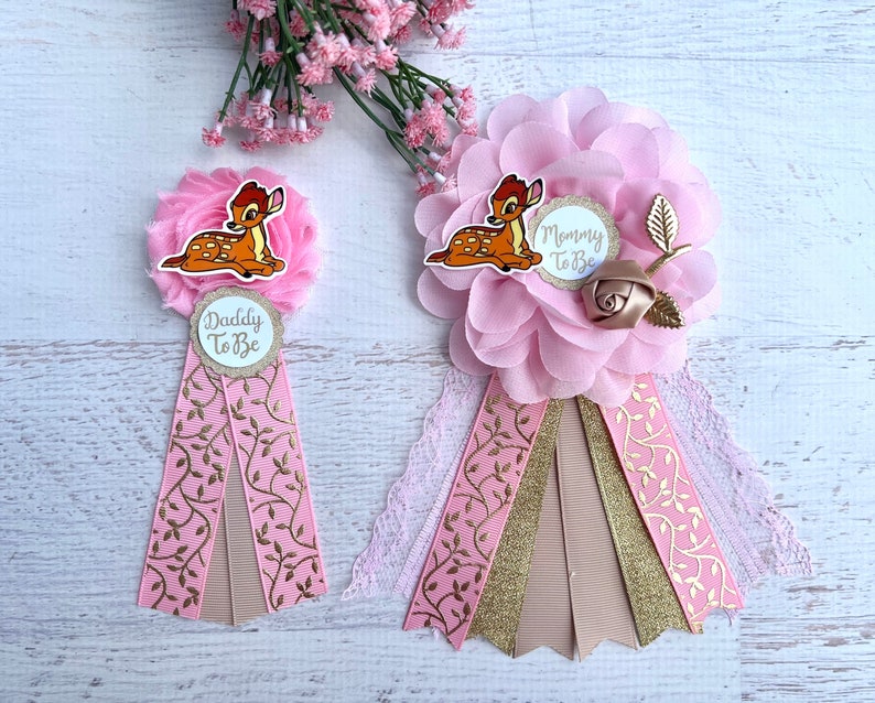 May include: Two pink and gold baby shower sashes with a cartoon Bambi design. The sashes have the text 'Mommy to Be' and 'Daddy to Be' on them