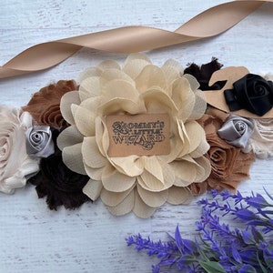 May include: A beige, brown, and black fabric flower sash with a brown tag that reads "Mommy's Little Wizard".