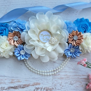 K&ouml;nnte beinhalten: Ein blaues und wei&szlig;es Satinsband mit einer gro&szlig;en wei&szlig;en Blume und einer kleineren blauen Blume. Das Band hat einen goldenen Kreis mit dem Text "Mommy To Be" in wei&szlig;. Das Band ist mit einer Perlenkette verziert.