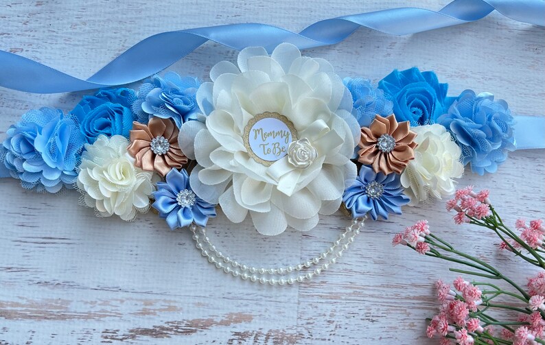 K&ouml;nnte beinhalten: Ein blaues und wei&szlig;es Satinsband mit einer gro&szlig;en wei&szlig;en Blume und einer kleineren blauen Blume. Das Band hat einen goldenen Kreis mit dem Text "Mommy To Be" in wei&szlig;. Das Band ist mit einer Perlenkette verziert.