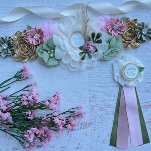 May include: A white satin sash with a floral arrangement in shades of pink, green, and gold. The sash has a white circle with the text "Mommy to Be" in gold lettering. A separate white flower with a gold circle and the text "Daddy to Be" is also included. The flower is attached to a green and pink ribbon.