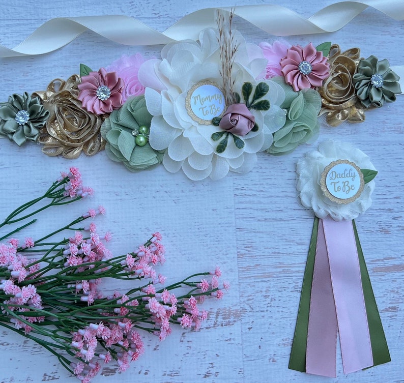 May include: A white satin sash with a floral arrangement in shades of pink, green, and gold. The sash has a white circle with the text "Mommy to Be" in gold lettering. A separate white flower with a gold circle and the text "Daddy to Be" is also included. The flower is attached to a green and pink ribbon.
