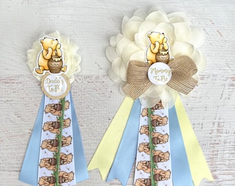 May include: Two Winnie the Pooh themed baby shower sashes. One sash is yellow and blue with the text "Mommy To Be". The other sash is blue and white with the text "Daddy To Be".