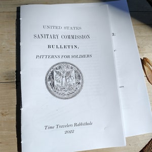 May include: A black and white image of a vintage book titled "United States Sanitary Commission Bulletin. Patterns for Soldiers". The book features a seal with the text "United States Sanitary Commission" and a design of a woman holding a staff and a shield. The book also includes the text "Time Travelers Rabbithole 2022".