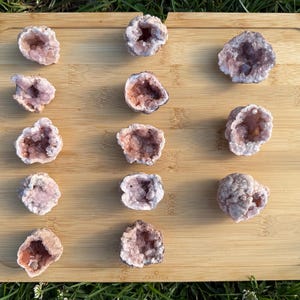 May include: A collection of fourteen small, rough-cut amethyst geode clusters arranged in a grid on a rectangular bamboo board. Each geode features a hollow center filled with light purple to pale pink crystalline structures and a textured exterior.
